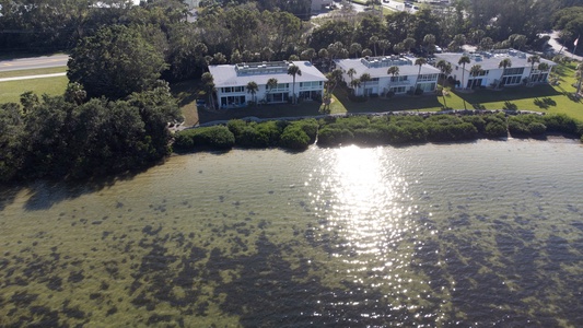 Aerial view of three modern waterfront houses with flat roofs, located next to a large body of water, surrounded by trees and a road in the background. Sunlight reflects off the water.