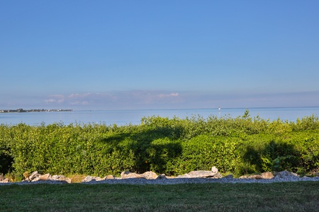 A serene coastal scene showing a grassy area beside a rocky shoreline, dense green shrubs, and a calm, blue sea under a clear sky.