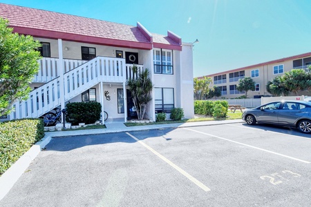 A two-story apartment building with outdoor stairs leading to the upper level. A bicycle is parked near the stairs. A car is parked in the parking lot in front, with designated parking spaces.