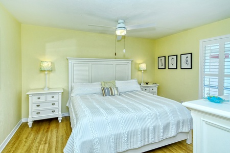 A bright bedroom with a large bed covered in a striped white blanket, flanked by two lamps on nightstands. Art hangs on the pale yellow walls, and a ceiling fan is above. Wooden floor and window with shutters.