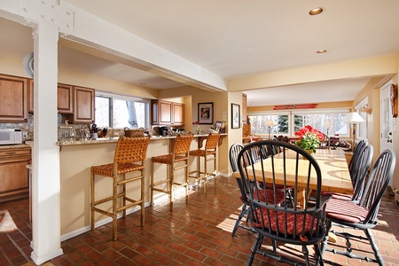 Open-plan dining area and kitchen with a wooden dining table, black chairs, a breakfast bar with wicker stools, and light wooden cabinets. The space features tile flooring and large windows.