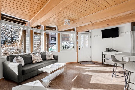 A cozy living room with a dark gray couch, glass coffee table, wall-mounted TV, and a small dining area. Large windows bring in natural light, highlighting the wooden ceiling and beams.