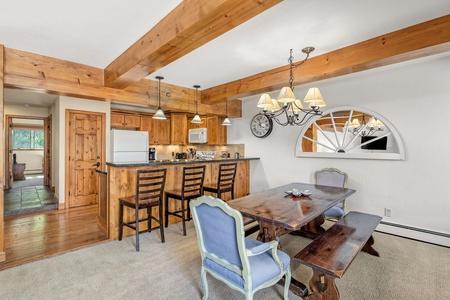 A rustic kitchen and dining area with wooden beams, a wooden dining table with mixed seating, a clock on the wall, and a partial view of the kitchen with bar stools at the counter.