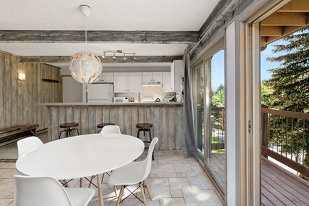 Modern kitchen and dining area featuring white round table with chairs, wooden beams, hanging light fixture, large window, and sliding door leading to outdoor balcony with trees visible.