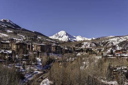 A snowy mountain landscape with buildings in the foreground, surrounded by bare trees and roads, under a clear blue sky.