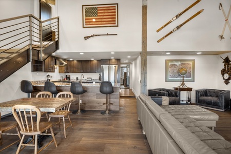 Modern living space featuring a kitchen with barstools, a dining table with wooden chairs, a leather couch, and wall-mounted decor including an American flag, rifle, and skis.