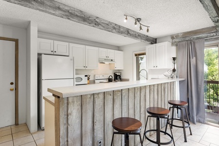 A kitchen with white cabinets, a white refrigerator, a wooden island counter, and three bar stools. There's a window with curtains and wooden beams on the ceiling.