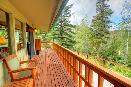 A wooden balcony with railings overlooks a forested area. The balcony has two chairs and a lamp near a large window and sliding door.