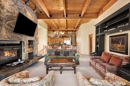 A cozy living room with a stone fireplace, mounted TV, and wooden ceiling beams. Furniture includes a green sofa, plaid sofa, and armchairs. The background shows a dining area and chandelier.