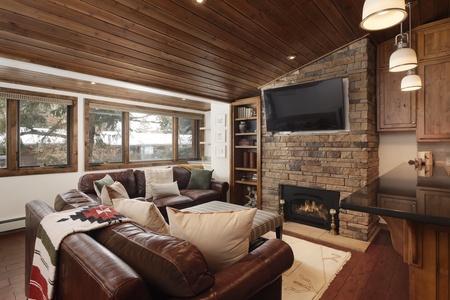 A cozy living room features a leather sectional, stone fireplace, wall-mounted TV, and large windows with a view of trees. Wood-paneled ceiling and warm lighting create a rustic ambiance.