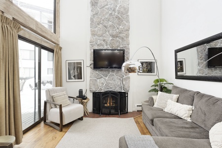 A cozy living room with a stone fireplace, wall-mounted TV, large mirror, grey sectional sofa, and modern floor lamp. Large windows and sliding glass door provide natural light.