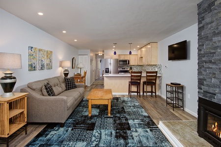 A modern living area with a grey couch, coffee table, and rug. The space features a stone fireplace, open kitchen with barstools, and a wall-mounted TV. Decor includes wall art and table lamps.