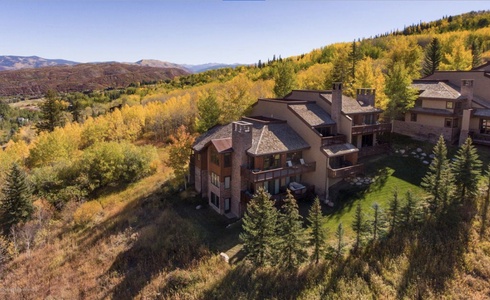 Large, multi-story house with balconies surrounded by trees with autumn foliage and mountainous background.