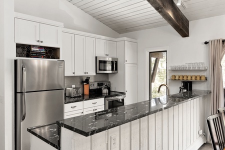 Modern kitchen with white cabinetry, stainless steel appliances, black granite countertops, and a white paneled island. The space features a vaulted ceiling with a wooden beam and a view of outdoors.