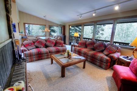 A cozy living room with large windows, two striped sofas, a glass coffee table, and a red armchair. The room features beige carpeting and a view of the trees outside.