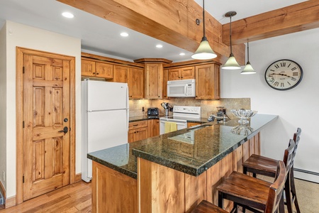 A kitchen with wooden cabinets and a green granite countertop, featuring a white refrigerator, oven, microwave, and bar seating. A wall clock is mounted above the countertop.