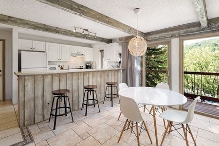 A modern kitchen with a rustic wooden bar featuring four stools, white cabinets, and a round dining table with four white chairs. Large windows and a door offer a view of greenery outside.