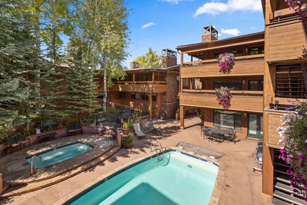 Outdoor view of a multi-story wooden lodge with balconies, surrounded by tall trees. A pool and hot tub are in the courtyard, with lounge chairs and potted flowers enhancing the scene.