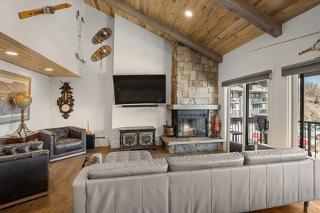 A cozy living room with a stone fireplace, mounted TV, gray sectional sofa, leather armchairs, and rustic decor, including a globe, clock, snowshoes, and skis. Large windows offer natural light.