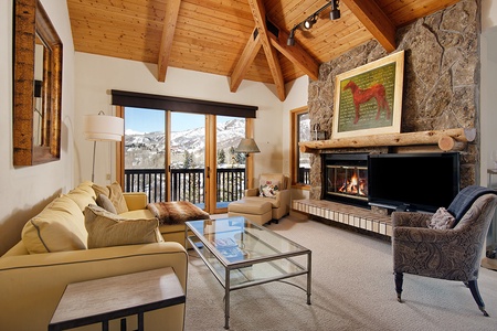 A cozy living room with vaulted wooden ceiling, large windows, stone fireplace, beige sofas, a glass coffee table, a patterned armchair, and a flat-screen TV. A painting of a horse hangs above the fireplace.