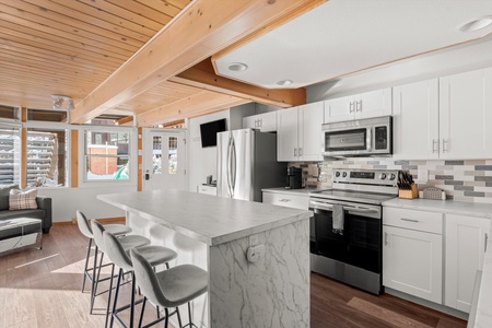 A modern kitchen with white cabinets, stainless steel appliances, a marble island with three gray stools, and wooden ceiling beams. The space is well-lit by natural light from large windows.