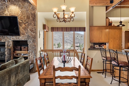 A dining room with a wooden table set for eight, adjacent to a kitchen with bar stools. There is a stone fireplace and a view of hills through large windows. An overhead light fixture illuminates the space.