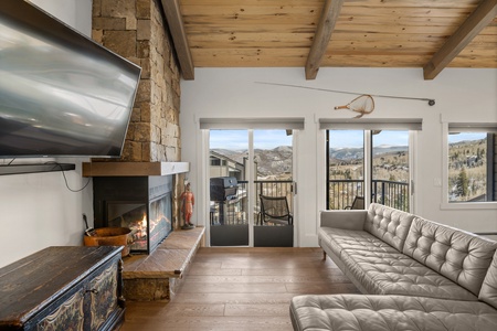 A modern living room with a wall-mounted TV, stone fireplace, gray sectional sofa, and large windows opening to a mountain view balcony. A rustic chest and fly fishing rod are also visible.