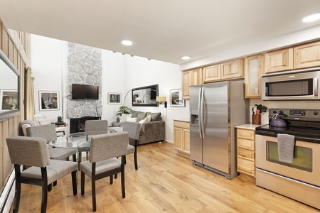 A modern kitchen with stainless steel appliances, light wood cabinetry, and an adjacent dining area with a glass table and gray chairs. A cozy living area with a sofa and flat-screen TV is visible in the background.