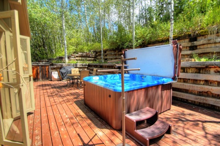 A hot tub with a blue cover partially open sits on a wooden deck. There are steps leading into the tub, and a seating area with a table and chairs is visible in the background.