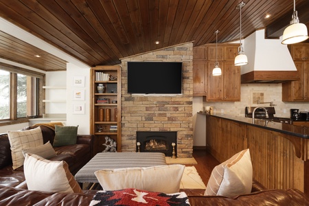 A cozy living area features a stone fireplace, mounted TV, and wooden ceiling. Leather couches and patterned cushions add comfort. Kitchenette with wooden cabinets is adjacent.