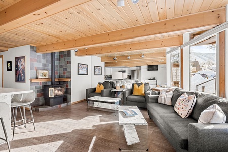 A modern living room with wooden ceiling beams, a fireplace, two gray sofas, and a large window offering a view of a snowy landscape. The space includes a kitchen with a white island and bar stools.