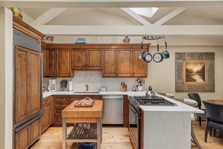 A modern kitchen with wooden cabinets, a stainless steel refrigerator, a kitchen island with a pot rack above, and a dining area. Artwork hangs on the wall beside the dining table with gray chairs.