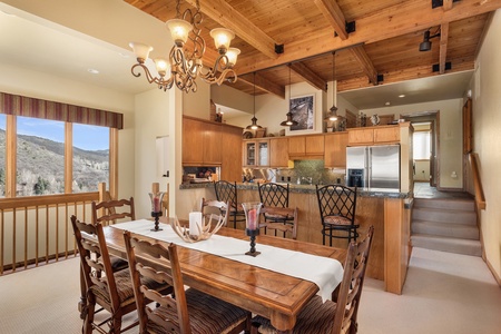 A wooden dining table with chairs is in the foreground, while a kitchen with stainless steel appliances and an island with barstools is in the background. The room has wooden ceilings and a window view.