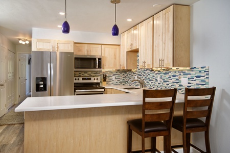 Modern kitchen with light wood cabinets, stainless steel appliances, mosaic tile backsplash, white countertops, two wooden chairs at the bar, and three pendant lights above.