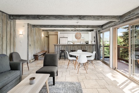 A bright living space with a gray sofa, white chairs around a dining table, a wooden coffee table, a kitchen with white cabinets, and large windows leading to a balcony with a view of greenery.