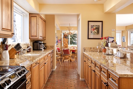 A kitchen featuring wooden cabinets, granite countertops, a stove, a coffee maker, utensils, and a sink. A dining area with chairs and a table is visible in the background.