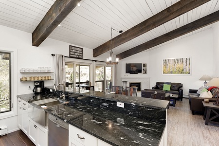 A modern kitchen with a black granite countertop, stainless steel appliances, and an open view into a living area with wooden beams on the ceiling and a fireplace.