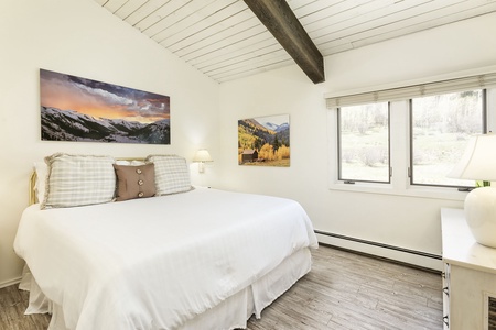 A bedroom with a white bed, wooden ceiling beam, landscape pictures on the walls, a window with blinds, and a bedside lamp on a white nightstand.