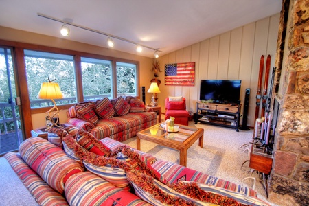 A cozy living room with striped furniture, a TV, a wooden coffee table, large windows, a flag on the wall, and decorative skis. A stone fireplace is partially visible to the right.