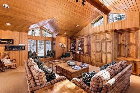 A cozy living room with wooden walls and ceilings, two plaid sofas, a coffee table, built-in bookshelves, and large windows offering a view of the snowy outdoors.