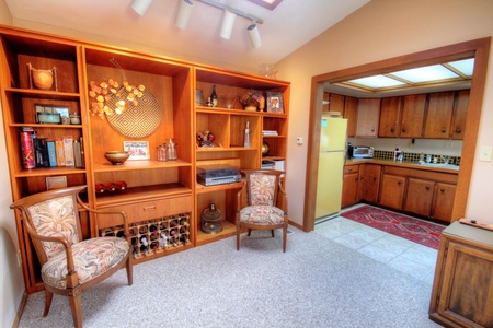 A room with wooden shelves displaying various items and two cushioned chairs. The shelves include books, decorations, and a wine rack. An open doorway leads to a kitchen with wooden cabinets and appliances.