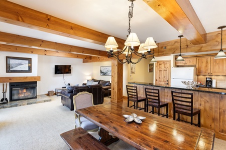 A cozy living area with wooden beams, a dining table, a fireplace, a wall-mounted TV, and a kitchen with wooden cabinets and black countertops. Three barstools are placed at the kitchen counter.