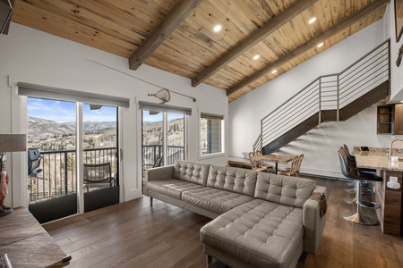 A modern living room with a large sectional sofa, open kitchen with bar stools, vaulted wooden ceiling, and a staircase. The room features a balcony overlooking a mountain landscape.