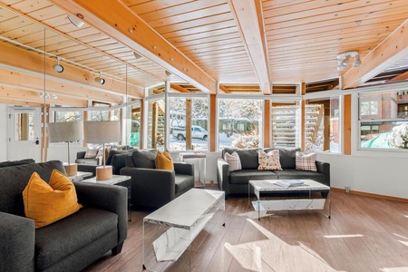 A modern living room with wooden ceiling, large windows, black sofas with yellow and patterned cushions, and two white marble-topped coffee tables on a wooden floor. Snowy landscape visible outside.