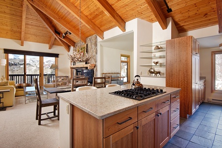 A modern kitchen and dining area featuring wooden cabinets, a gas stove on a central island, a stone fireplace, and large windows with mountain views. The space has a high wooden ceiling with exposed beams.