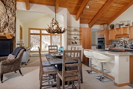 A cozy open-plan kitchen and dining area with a wooden table and chairs, a white kitchen island with stools, a stone fireplace, and a ceiling with exposed wooden beams.