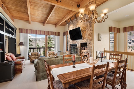 A cozy living and dining room with wooden beams on the ceiling, a stone fireplace, a wall-mounted TV, large windows, and a dining table with chairs. The room has a view of trees and mountains outside.