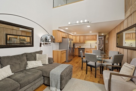 A cozy living room with a gray sectional sofa, glass-top dining table, modern kitchen with wooden cabinets, reflective mirrors, and hardwood floors.