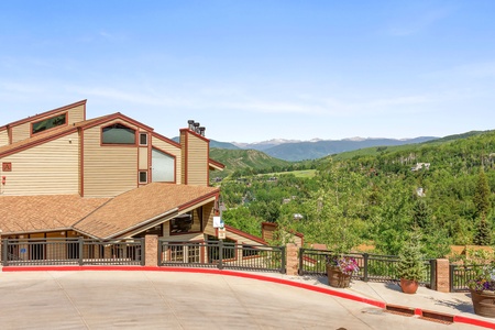 A wooden house with a pitched roof, large windows, and chimney on a mountainous landscape with green trees and distant peaks under a clear blue sky.