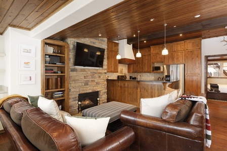 A cozy living room with leather sofas, cushions, and a fireplace. The room features wooden ceilings and cabinetry, a mounted TV, and pendant lighting. An adjacent kitchen with stainless steel appliances is visible.